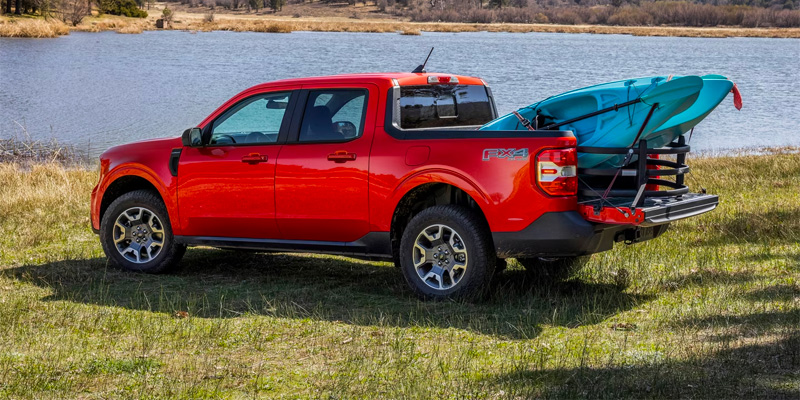 A red 2025 Ford Maverick parked on grass next to a lake with a kayak in the truck bed