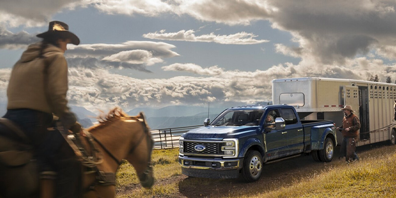 A man on a horse facing a blue 2025 Ford F-250 Super Duty pickup truck that has a trailer in tow and parked next to a lake with a slightly cloudy blue sky overhead