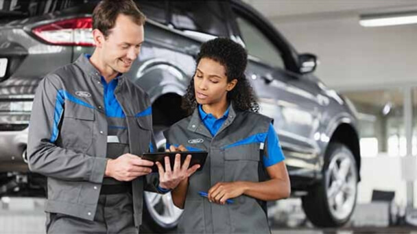 Two Ford Certified Service Technicians reviewing details on a tablet for a service customer with a grey Ford vehicle in the background
